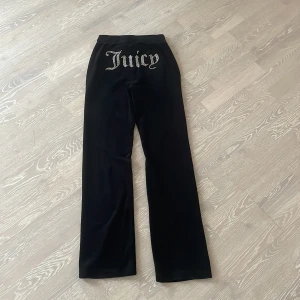Svarta mjukisbyxor från Juicy Couture - Snygga svarta mjukisbyxor från Juicy Couture med glittrig text på baksidan. Byxorna har en bekväm passform med snörning i midjan och ett litet JC-emblem framtill.Jag säljer pga att jag har vuxit ur dom.Vra skick förutom att det saknas en plupp på snöret i midjan