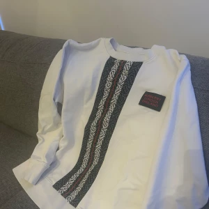 Vit långärmad tröja från Burberry - Säljer en stilren vit långärmad tröja från Burberry med ett unikt mönster i svart och rött längs framsidan. Tröjan har en klassisk passform och ett märke med texten 'Burberry London England' på bröstet.