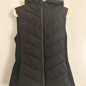 Svart dunväst från Zara - Snygg svart dunväst från Zara med dragkedja och huva. Perfekt för kyliga dagar när du vill hålla värmen utan att kompromissa med stilen. Västen har en quiltad design som ger en modern look.