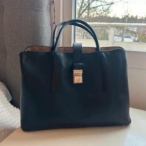 Svart handväska med låsdetalj - Elegant svart handväska i skinn med en stilren design. Väskan har två handtag och en snygg låsdetalj i metall på framsidan. Perfekt för att bära dina dagliga nödvändigheter med stil.