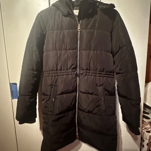 Svart dunjacka med huva - Snygg svart dunjacka med huva och dragkedja framtill. Jackan har långa ärmar och praktiska fickor med knappar. Perfekt för kyliga dagar. Mycket välvårdad. Till barn str 158
