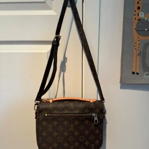 Brun axelväska med monogram - Snygg brun axelväska med klassiskt monogrammönster och gulddetaljer. Väskan har en justerbar axelrem och en praktisk dragkedja framtill. Insidan är rymlig med flera fack för enkel organisering. Perfekt för att bära med sig det viktigaste med stil.