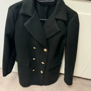 Svart kappa med knappar - Elegant svart kappa med dubbelknäppning och breda slag. Perfekt för en stilren look. Kappan har en klassisk design med långa ärmar och dekorativa knappar framtill.