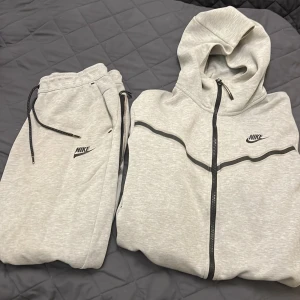 Grå nike tech hoodie och Grå Nike tech byxor för endast 600 kr  - Säljer en grå hoodie från Nike med dragkedja och svart logotyp på bröstet. Den har en justerbar huva med snören och är perfekt för en avslappnad stil.