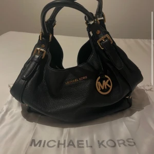 Svart skinnväska från Michael Kors - Snygg svart skinnväska från Michael Kors med gulddetaljer och logotyp. Väskan har dubbla handtag och en praktisk dragkedja. Perfekt för att bära med sig alla nödvändigheter med stil.
