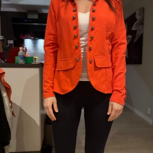 Orange kavaj med knappar - Snygg orange kavaj med dekorativa knappar framtill och på ärmarna. Den har en figurnära passform och två fickor fram. Perfekt för att liva upp garderoben med en färgklick! Stolek S. Ifrån Copenhagen. 