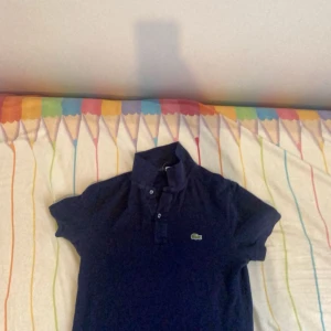 Mörkblå pikétröja från Lacoste - Säljer en snygg mörkblå pikétröja från Lacoste. Den har korta ärmar och den klassiska krokodilloggan på bröstet. Perfekt för en avslappnad men stilren look!, har andvänts få gånger! Köpt för ungefär 2 månader sen, pris kan diskuteras! :)
