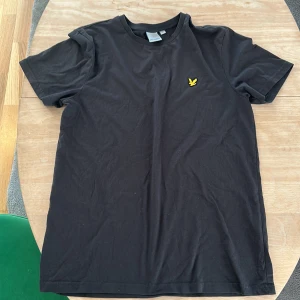Svart t-shirt från Lyle & Scott - Säljer en klassisk svart t-shirt från Lyle & Scott med deras ikoniska gula logga på bröstet. T-shirten är i storlek L och har en normal passform. Perfekt för vardagsbruk eller en avslappnad stil.