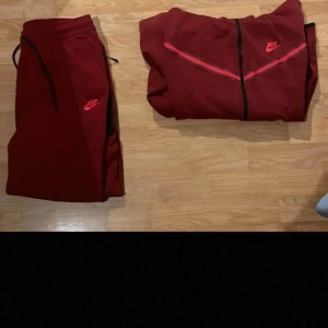 Röd tech dress från Nike - Säljer en snygg röd hoodie och byxor från Nike. Den har en dragkedja framtill och en cool design med svarta detaljer. Perfekt för höst och vinter, och passar både till träning och vardag. Nike-loggan syns tydligt på bröstet. Superbekväm och stilren!