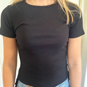 Svart ribbad topp - Säljer en svart ribbad topp med korta ärmar. Den är croppad och har en snygg knytning i sidan som ger en extra detalj.