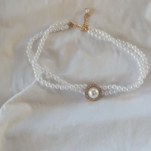 Dubbelradigt pärlhalsband med gulddetaljer - Dubbelradigt pärlhalsband med en stor pärla i mitten omgiven av gnistrande stenar. Halsbandet har guldfärgade detaljer och en justerbar kedja för perfekt passform. Perfekt för festliga tillfällen eller som en lyxig accessoar till vardagsoutfiten.