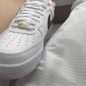 Nike Air Force 1 '07 LV8 i vitt och svart - Säljer ett par Nike Air Force 1 '07 LV8 sneakers i klassisk vit färg med svart swoosh. Skorna har snygga gulddetaljer vid snörningen och är tillverkade i skinn. Perfekta för både vardag och fest!