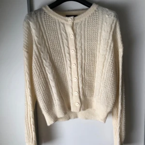 Vit stickad kofta från Cider - Säljer en superfin vit stickad kofta från Cider. Den har ett klassiskt flätmönster och knäpps med söta vita knappar framtill. Perfekt för höst och vinter när du vill hålla dig varm och stilren. Koftan är långärmad och sitter som tshirt. Knappt använd