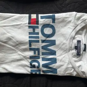 Säljer en vit t-shirt från Tommy Hilfiger med stort blått tryck på framsidan. T-shirten är i bomull och har korta ärmar. Perfekt för en avslappnad stil. Den är lite skrynklig men i bra skick.