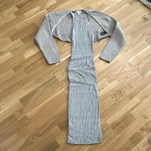 Randig långklänning med bolero - Snygg randig långklänning i ljusgrått och vitt med matchande bolero. Klänningen är ärmlös och har en tight passform som framhäver figuren. Perfekt för en stilren look.