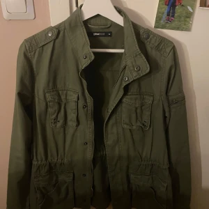 Grön jacka från Gina Tricot - Säljer en snygg grön jacka från Gina Tricot. Jackan har långa ärmar och flera fickor framtill med knappdetaljer. Perfekt för höst och vår med sin stilrena design och bekväma passform.