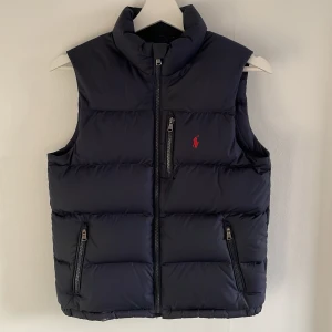 Blå dunväst från Ralph Lauren - Säljer en stilren blå dunväst från Ralph Lauren med dragkedja och hög krage. Västen har tre fickor med dragkedjor och en liten röd logotyp på bröstet. Perfekt för kyligare dagar.                         Svarar snabbt på frågor!