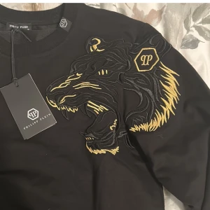 Svart tröja med lejonbroderi från Philipp Plein - Snabb köp=bra pris!! Snygg svart tröja från Philipp Plein med ett stort, detaljerat lejonbroderi i guld på bröstet. Tröjan har långa ärmar och ribbade muddar. Perfekt för en stilren look.