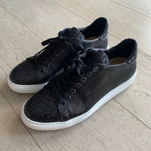 Svarta sneakers med flätad detalj CC Shoes - Snygga svarta sneakers med flätad detalj på tå och häl. Skorna har en vit sula och snörning framtill. Perfekta för en stilren look. Handgjorda i Italien av äkta kalvskinn. Använda bara en gång. Storleken är 41 1/2 men passar även 42 och 42 1/2. Ordinarie pris 4500kr