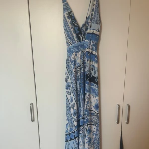 Blå och vit långklänning - Snygg långklänning i blått och vitt med ett unikt mönster. Klänningen har en v-ringad design och är ärmlös, vilket ger en elegant look. Perfekt för sommarens alla tillfällen.