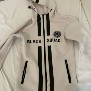 Vit jacka med tryck - Säljer en vit jacka med texten 'BLACK SQUAD' och ett tryck på bröstet. Jackan har en dragkedja framtill och två fickor med dragkedjor. Den har en huva med dragsko för justering.
