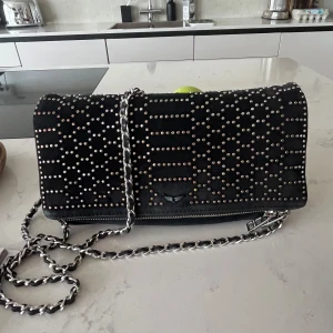 Zadig et Voltaire Svart axelväska med nitar - Snygg svart axelväska dekorerad med små nitar över hela ytan. Väskan har en kedjerem som ger en cool touch och en dragkedja för säker förvaring. Perfekt för att ge din outfit lite edge. Lite missfärgning på några nitar men syns inte så mycket. Pris kan diskuteras 