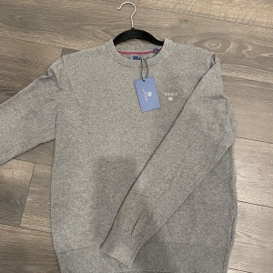 Grå Gant tröja i kashmir - Säljer en stilren grå tröja från GANT med broderad logotyp på bröstet. Tröjan har långa ärmar och ribbade muddar vid ärmslut och nederkant. Perfekt för en avslappnad och snygg look.