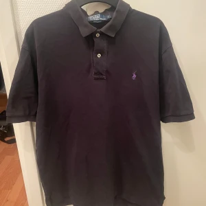 Svart pikétröja från Ralph Lauren - Säljer en klassisk svart pikétröja från Ralph Lauren. Tröjan har korta ärmar och en liten broderad logotyp på bröstet. Perfekt för en stilren look. Hör av dig vid intresse!