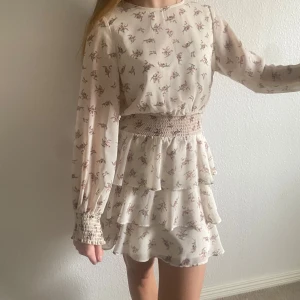 Blommig klänning med volanger från Gina Tricot  - Charmig blommig klänning i ljus färg med långa ärmar och volanger. Den har en smockad midja och en liten öppning i ryggen. Perfekt för en romantisk look. Säljer denna jättefina volang klänning från Gina Tricot då den inte kommer till användning. Fint skick då den är sparsamt använd. Perfekt för vardag eller fest! Skriv för fler bilder eller frågor!💕
