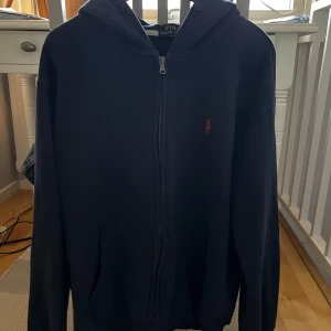 Mörkblå hoodie från Polo Ralph Lauren - Säljer en mörkblå hoodie från Polo Ralph Lauren med dragkedja och huva. Den har en röd broderad logotyp på bröstet och är i en lite mindre passform då den krympt i tvätten. Perfekt för en avslappnad stil. Storlek S nu men var en M.