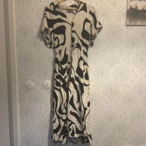 Svartvit mönstrad klänning - Säljer en lång svartvit klänning med abstrakt mönster. Klänningen har korta ärmar och ett knytband i midjan för en justerbar passform. Perfekt för en stilfull look.