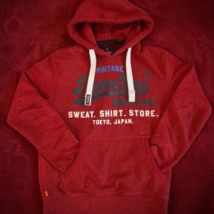 Röd hoodie från Superdry - Säljer en snygg röd hoodie från Superdry med tryck i blått och vitt. Hoodien är sparsamt använd och har inga defekter.