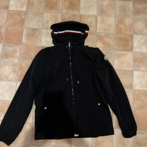 Svart moncler vindjacka med huva - Snygg svart moncler vindjacka med huva och dragkedja framtill. Jackan har långa ärmar och en stilren design med ränder på huvan. Perfekt för blåsiga dagar. Storlek Xl men sitter som L