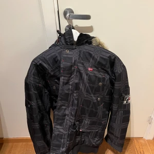 Vinter Jacka  Från Geographical Norway - Säljer en svart rutig jacka från Geographical Norway med pälsfodrad huva. Jackan har dragkedja och knappar framtill samt flera praktiska fickor. Perfekt för kyliga dagar med sin quiltade insida och stilrena design.  Jackan är aldrig använd och storlek XL