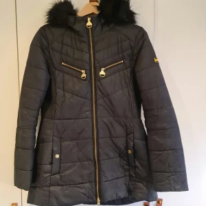 Svart quiltad jacka från Barbour - Barbour Grounding Quilt lättviktsjacka med fuskpäls på huvan och fina guldfärgade dragkedjor och knappar. Superskön, figurformad och väger ingenting. Gjord av fibre down som håller värmen. Perfekt under kallare höst/vår eller mildare vinter. Jackan har två bröstfickor med dragkedja och två sidofickor framtill. En inneficka. En knapp har lossnat på ena sidofickan där revan är lagad med specialtejp för just denna typ av jackor (se bild). Sparsamt använd.
