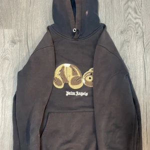 Svart hoodie från Palm Angels - Säljer en svart hoodie från Palm Angels med ett unikt nallemotiv på framsidan. Hoodien har en klassisk passform med känguruficka och justerbar huva. Tillverkad i Italien, perfekt för en stilren och avslappnad look.