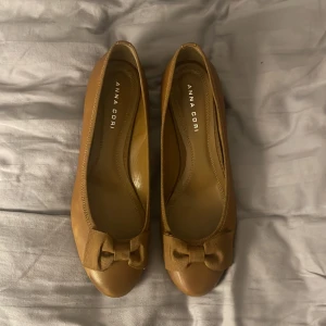 Bruna ballerinaskor från Anna Cori - Säljer ett par eleganta bruna ballerinaskor från Anna Cori. Skorna har en söt rosett framtill och är tillverkade i skinn. Perfekta för en stilren look.