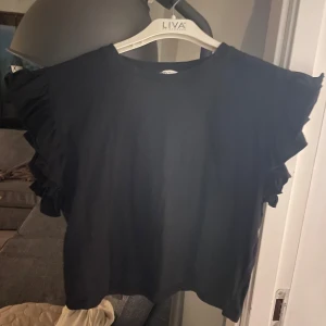 Svart topp med volangärmar från Zara - Säljer en svart topp från Zara med volangdetaljer på ärmarna. Toppen har en rund hals och är perfekt för en stilren look. Passar bra till både jeans och kjol.