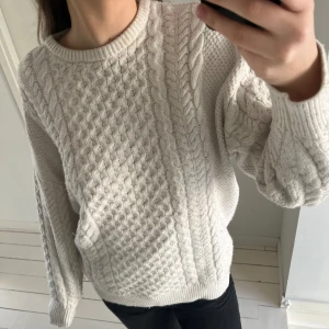 Beige stickad tröja - Mysig beige stickad tröja med kabelstickat mönster. Tröjan har långa ärmar och en rund halsringning, perfekt för kyligare dagar. Den är både stilren och bekväm, vilket gör den till ett mångsidigt tillskott i garderoben.