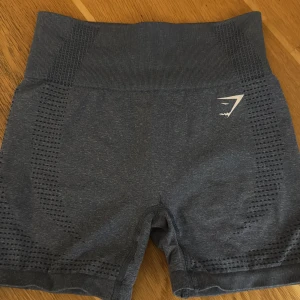 Gråblå shorts från Gymshark - Säljer ett par gråblå shorts från Gymshark med hög midja och tight passform. Perfekta för träning med sitt stretchiga och bekväma material. De har en diskret logga på sidan och ett mönster som ger extra ventilation.