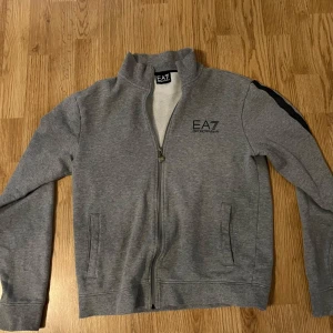 Grå zip-tröja från EA7 - Snygg grå zip-tröja från EA7 med svart logga på bröstet och svarta detaljer längs ärmarna. Tröjan har en dragkedja framtill och är tillverkad i ett mjukt material. Perfekt för en casual look.