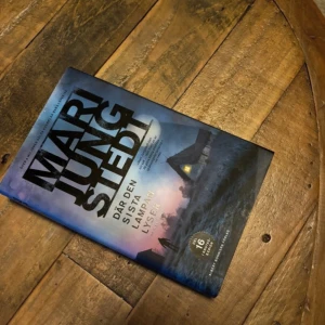 Där den sista lampan lyser - Boken 'Där den sista lampan lyser' av Mari Jungstedt är en spännande deckare. Omslaget visar en mystisk kustscen i skymningen med en ensam stuga vars fönster lyser. Perfekt för dig som älskar kriminalromaner och vill ha något fängslande att läsa.