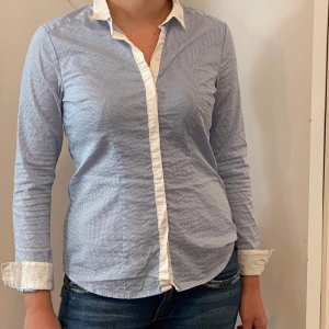 Blå och vit randig skjorta - Säljer en stilren blå och vit randig skjorta i storlek 40. Skjortan har långa ärmar och en klassisk krage med vita detaljer. Perfekt för både vardag och jobb. Den är i bra skick och gjord av ett bekvämt material. Passar perfekt till jeans för en avslappnad look.