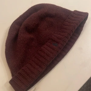 Vinröd mössa från Polo Ralph Lauren - Säljer en snygg vinröd mössa från Polo Ralph Lauren. Den är stickad och perfekt för höst och vinter. Mössan har en ribbad kant som ger en skön passform. Den är i nyskick och redo att hålla dig varm och stilren i kylan. Passar perfekt till både vardag och lite finare tillfällen.