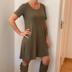 Olivgrön klänning från Cotton Sense - Säljer en superfin olivgrön klänning från Cotton Sense. Den är kortärmad och har en avslappnad passform som är perfekt för både vardag och sommar. Materialet är mjukt och bekvämt, vilket gör den idealisk för varma dagar. Perfekt att styla med stövlar eller sneakers!