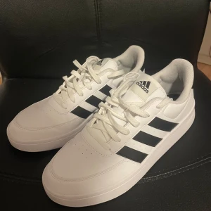 Vita Adidas sneakers med svarta detaljer - Säljer ett par snygga vita Adidas sneakers i nyskick. De har de klassiska tre svarta ränderna på sidan och Adidas-loggan på plösen och hälen. Perfekta för både vardag och träning. Skorna har snörning och är i storlek herr 44. Passar perfekt till alla outfits!