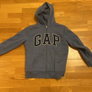 Blå hoodie från GAP - Säljer en supermysig blå hoodie från GAP. Den har en stor logga på framsidan och en dragkedja hela vägen ner. Insidan är fodrad med mjuk fleece, perfekt för kyliga dagar. Den har långa ärmar och en praktisk huva. Perfekt för både vardag och chill!