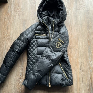 Svart quiltad jacka från Rockandblue - Säljer en snygg svart quiltad jacka från Rockandblue. Den har en cool design med guldiga dragkedjor och ett märke på ärmen. Perfekt för att hålla sig varm och stilren under kalla dagar. Jackan har en huva och flera praktiska fickor.