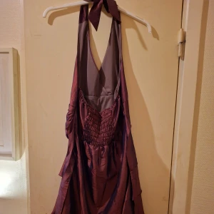 Vinröd klänning strl S - En vacker klänning i en mörkare röd färg med en halterneck och en rosett i ryggen. Klänningen har en flerskiktad kjol och spets längst ner. Använd en gång.