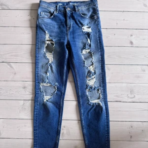 Blå jeans med slitningar - Säljer ett par snygga blå jeans med coola slitningar på framsidan. De har en klassisk femficksdesign och en knappgylf. Perfekta för en avslappnad och trendig look!